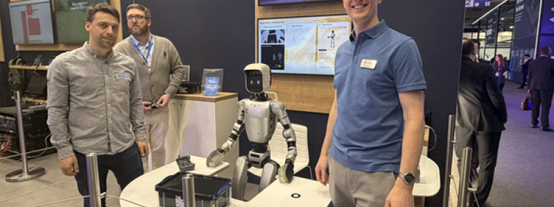 Dom Rugg-Gunn (till höger), robotikingenjör på Cambridge Consultants, vid bolagets monter på MWC 2026. Den humanoida roboten mellan teammedlemmarna är den som under mässan demonstrerade hur AI kan tolka mänskliga gester och plocka upp objekt. Foto: Nicholas Christiansen.